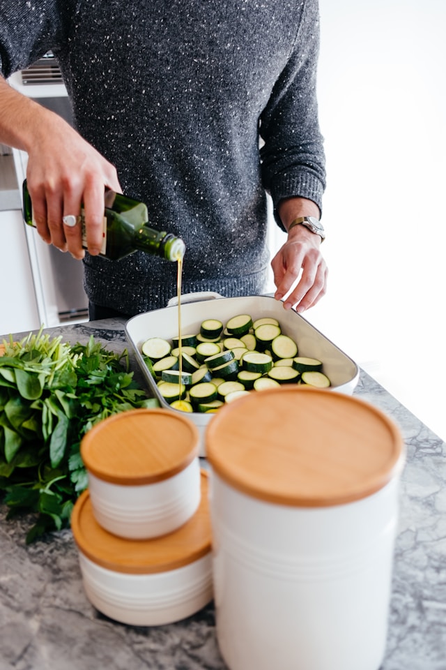 Person in a home kitchen preparing a salad, pouring olive oil into a bowl with salad ingredients on the table and inside the bowl.