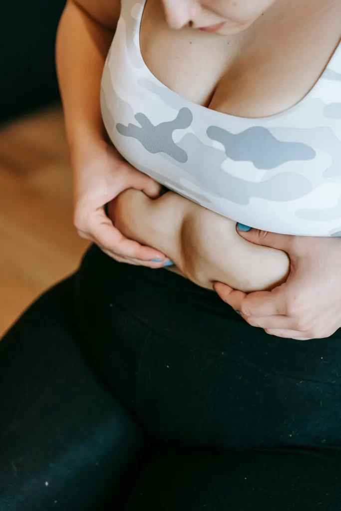Young overweight woman sitting, holding excess belly fat with both hands, wearing gym clothes, looking sadly at her stomach.